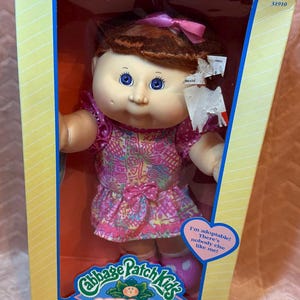 Peut inclure: Une poupée Cabbage Patch Kids aux cheveux auburn avec un nœud rose, portant une robe rose à motifs avec un petit nœud à la taille. La poupée est dans une boîte jaune et bleue avec le texte Cabbage Patch Kids, Kids, et I'm adoptable!