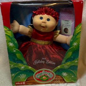 May include: A 2018 Holiday Edition Cabbage Patch Kids doll with blue eyes and dark hair, wearing a red satin dress with gold trim and a matching red floral headband. The doll is packaged in a box featuring green and white snowflake graphics.