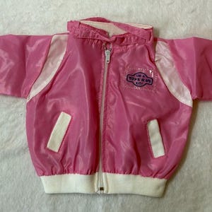 May include: A pink vintage jacket with white detailing and a full zip. The jacket features a small patch that reads "I'M A TOYS R US KID!". The cuffs and hem are white.