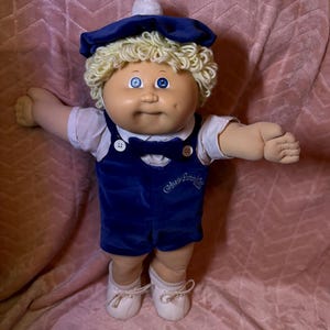 Op de afbeelding: Een Cabbage Patch Kids pop met blond garen haar, blauwe ogen en een gezicht met kuiltjes. De pop draagt een wit overhemd met kraag, een donkerblauwe fluwelen tuinbroek met bijpassende vlinderdas, een donkerblauwe baret en witte schoenen.