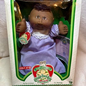Peut inclure: Une poupée Cabbage Patch Kids Preemie à la peau foncée et aux cheveux bruns, vêtue d'une robe violet clair avec un col en dentelle blanche. La poupée est dans une boîte avec la marque 25th Anniversary, un certificat de naissance et le texte Only at Target.
