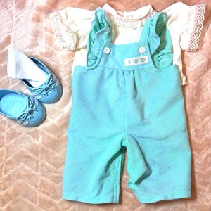 May include: A set of doll clothing consisting of a pair of light turquoise corduroy overalls with ruffled straps and two white buttons, a cream-colored short-sleeved top with pink lace trim on the sleeves, and two light blue doll shoes.