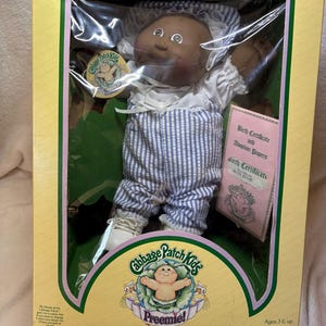 Pode incluir: Uma boneca Cabbage Patch Kids Preemie de pele escura, vestindo uma camisa branca e jardineiras às riscas azuis e brancas com um gorro a condizer. A boneca está dentro de uma caixa de cartão amarela e verde com certidão de nascimento.