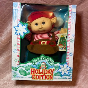 May include: A Cabbage Patch Kids Holiday Edition doll in its original packaging. The doll wears a red hat, a red and white striped shirt, and a brown vest with a gold buckle. The box features text including To, From, Cabbage Patch Kids, Holiday Edition, and Exclusively at K-MART.