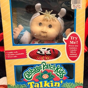 May include: A Cabbage Patch Kids Talkin' Farm Babies doll in a box. The doll is wearing a cow costume. The box has text that says "Try Me! Press my tummy, I make REAL animal sounds!" and "Cuddly soft. Machine washable. Dryer safe."
