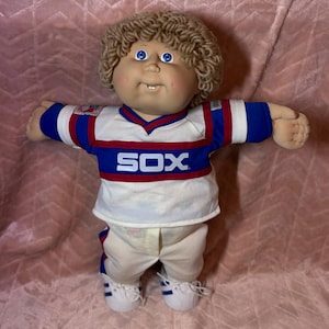 May include: A doll with light brown yarn hair and blue eyes wearing a white, blue, and red jersey with the text SOX printed on the chest. The doll is dressed in matching white trousers with blue and red side stripes and white athletic shoes.