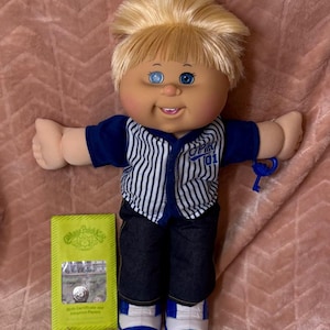 May include: A Cabbage Patch Kids doll with blonde hair and blue eyes, wearing a blue and white striped baseball-style shirt with CPK 01 text, dark blue trousers, and blue and white trainers. A small blue key accessory and a green adoption paper envelope.