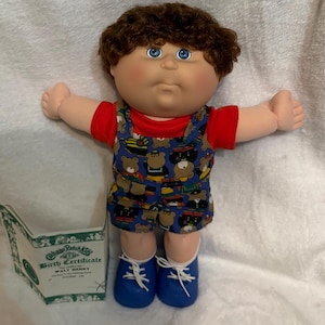 May include: A Cabbage Patch Kids doll with brown curly hair, blue eyes, and a red shirt. The doll wears blue overalls patterned with teddy bears and blue shoes. A birth certificate is visible. The doll's name is Walt Henry.