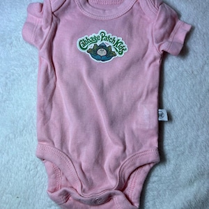 May include: A light pink short-sleeved bodysuit featuring a round neckline and a graphic print on the chest. The graphic displays the text Cabbage Patch Kids in green letters above an illustration of a doll emerging from a green cabbage leaf.