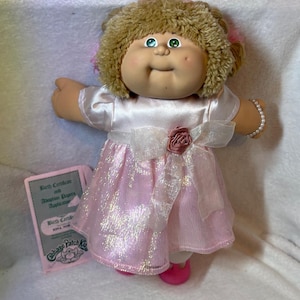 May include: A Cabbage Patch Kids doll wearing a pink dress with a white bow and a pearl bracelet. The doll has blonde hair and green eyes. A birth certificate and adoption papers are included.