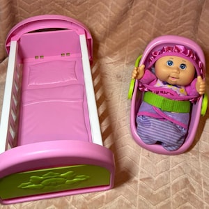 May include: A pink and white plastic doll crib with a lime green accent panel featuring a sun design, paired with a pink plastic doll carrier containing a doll with blue eyes, wearing a purple and striped outfit with a matching ruffled bonnet.
