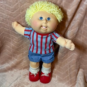 May include: A Cabbage Patch Kids doll with bright yellow yarn hair, blue eyes, and a smiling expression. The doll wears a red and blue striped short-sleeve shirt with a small CPK logo, blue denim shorts, white socks, and red shoes with white laces.