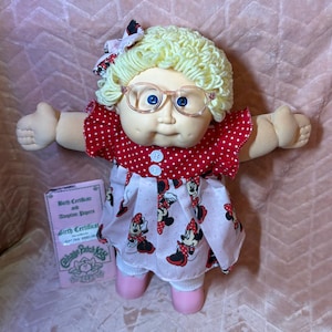 Könnte beinhalten: Eine Cabbage Patch Kids Puppe mit blondem Wollhaar, blauen Augen und einer transparenten Brille. Die Puppe trägt ein rotes Kleid mit weißen Punkten und Minnie Maus Motiv, eine passende Haarschleife und rosa Schuhe. Eine Geburtsurkunde und Adoptionspapiere liegen bei.
