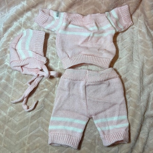 May include: A three-piece knitted set consisting of a short-sleeved off-the-shoulder top, shorts, and a bonnet. Each piece is crafted in a soft pastel pink yarn featuring horizontal white stripes and ribbed cuffs at the sleeves, waist, and leg openings.