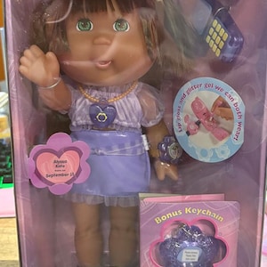 May include: A Cabbage Patch Kids Pretty Surprise doll named Alyssa Kate with brown hair in pigtails, wearing a lavender top and skirt. The packaging includes a bonus purple glittery star keychain, a toy phone, and lip gloss and glitter gel net wt 1.0 oz.