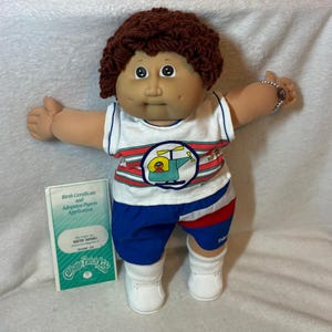May include: A Cabbage Patch Kids doll with brown yarn hair, dimpled cheeks, and brown eyes. The doll wears a white sleeveless top with a helicopter graphic, blue shorts with red accents, and white shoes. A birth certificate and adoption papers are included.
