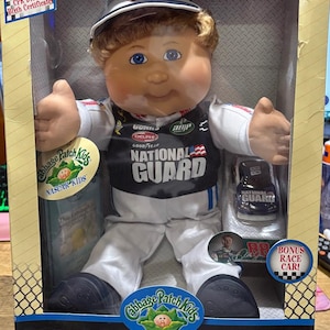 May include: Cabbage Patch Kids NASCAR doll in a clear box. The doll wears a racing uniform with "NATIONAL GUARD" on the front. Includes a bonus race car and a CPK Driver's Birth Certificate. The box has the NASCAR Kids logo.