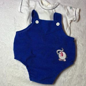 May include: A blue corduroy romper with white buttons and an embroidered elephant design, paired with a white short-sleeved shirt. The shirt has a black stitched neckline and sleeve hems. A vintage baby outfit.