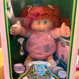 May include: A Cabbage Patch Kids doll in a box. The doll has red yarn hair, blue eyes, and is wearing a pink overall. The box has the text "25th Anniversary" and "Limited Edition". A silver spoon is included.