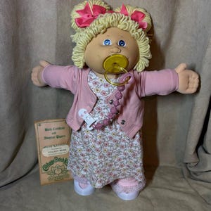 Può includere: Una bambola Cabbage Patch Kids con capelli in lana bionda a codini, occhi blu e un ciuccio giallo. La bambola indossa un cardigan rosa sopra un vestito a fiori con una catenella portaciuccio e scarpe bianche. Include certificato di nascita.
