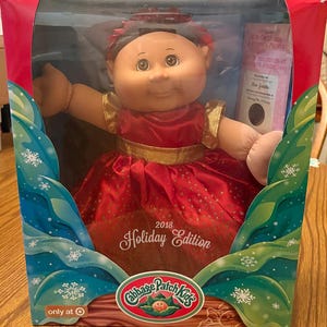 May include: A Cabbage Patch Kids 2018 Holiday Edition doll in a box. The doll wears a red dress with gold accents. The box has a festive design with snowflakes and the Target logo.