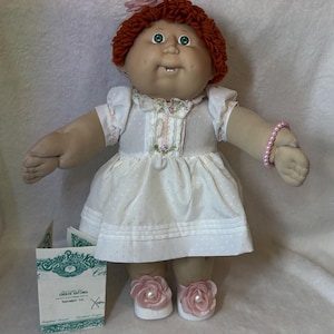 Peut inclure: Une poupée Cabbage Patch Kids avec des cheveux en laine auburn, des yeux verts et une seule dent. La poupée porte une robe blanche à pois discrets, une bordure en dentelle et un accessoire de cheveux fleur rose. Un bracelet de perles roses et des chaussures blanches avec des fleurs roses sont visibles.