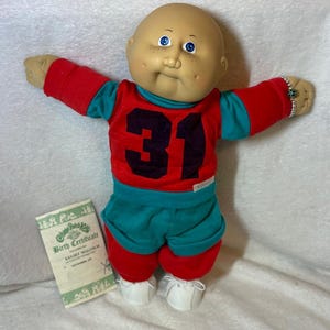 May include: A Cabbage Patch Kid doll wearing a red jersey with the number 31, teal shorts, and white shoes. The doll has a bald head, blue eyes, and a birth certificate that reads "Kelsey Malcolm". A silver bracelet with a green gem is on the doll's wrist.