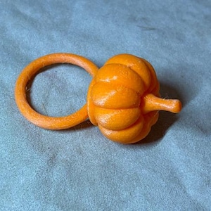 May include: A bright orange ring featuring a small, textured pumpkin charm attached to one side. The pumpkin has a rounded, ribbed shape with a short stem extending outward. The entire object is a uniform, vibrant orange color with a matte finish.