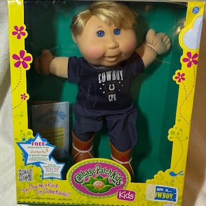 May include: A Cabbage Patch Kids doll with blonde hair and blue eyes, wearing a navy blue t-shirt with the text COWBOY CPK and a horseshoe graphic, dark trousers, and brown boots. The doll is packaged in a yellow and green box with floral accents.