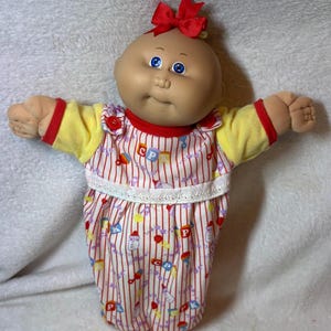 May include: A doll with a round head, blue eyes, and a red bow on top. It wears a yellow shirt with red trim and a white romper featuring red vertical stripes, baby bottle graphics, and blocks with the letters CP and K printed on the fabric.
