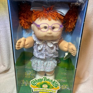 May include: A Cabbage Patch Kids doll with orange yarn hair in pigtails, wearing light purple glasses, a white collared shirt with a blue pattern, and a matching skirt. The doll is inside a blue box with a green field graphic and text.
