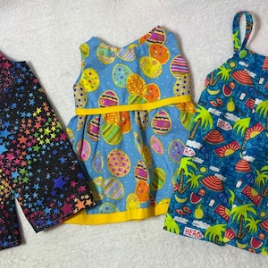 Pode incluir: Três vestidos de boneca coloridos. Um é preto com um padrão de estrelas arco-íris, outro é azul com desenhos de ovos de Páscoa, e o terceiro é azul com um tema de praia. Todos são sem mangas.