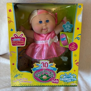 May include: A Cabbage Patch Kids doll with blonde hair featuring pink streaks, blue eyes, and a pink satin dress with a white bow. The doll is packaged in a yellow box with text including Celebrating 30 Years, 1983-2013, and a hair comb accessory.