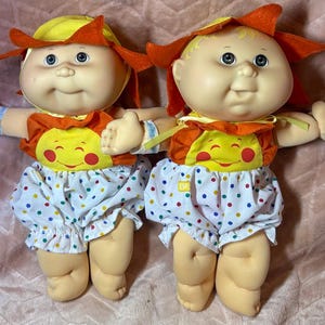 May include: Two dolls with light skin, blue eyes, and blonde hair, wearing orange sun-shaped hats and matching tops with a yellow sun graphic. They wear white bloomers with colorful polka dots and have light blue hospital-style wristbands on.