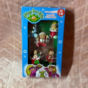May include: A Cabbage Patch Kids ornament set in a blue rectangular box featuring four small doll figures. The figures include a baby in a blue outfit, a baby in a red outfit, a baby in a red stocking, and a baby in a green outfit. The box displays the text Cabbage Patch Kids, Ornament Set, and Ensemble d'Ornaments.