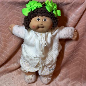 May include: A doll with dark brown yarn hair styled in two pigtails, each adorned with a bright lime green ribbon bow. The doll has a tan complexion, rosy cheeks, and wears a white knitted cardigan over white shorts with a yellow floral pattern.