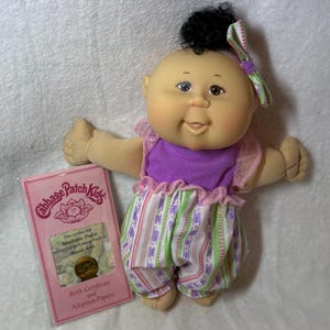May include: A Cabbage Patch Kids doll, Madison Paris, with black curly hair and a green and purple bow. The doll wears a purple top and striped pants. A pink birth certificate and adoption papers are included.