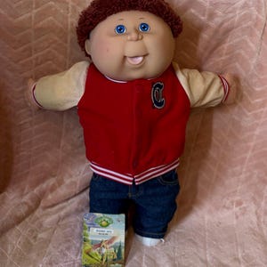 May include: A doll with brown curly hair and blue eyes wearing a red and cream varsity jacket with a letter C on the chest, paired with dark blue denim jeans. A small rectangular card with a graphic illustration and text is positioned in front.