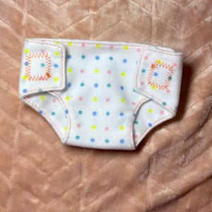 May include: A white fabric diaper cover featuring a repeating pattern of small blue, yellow, and pink polka dots. The garment includes two side tabs secured with orange zigzag stitching along the edges and orange trim around the leg openings.