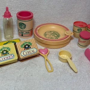May include: A collection of Cabbage Patch Kids toy accessories including two baby bottles, a canister, a plate, three small containers, two rectangular boxes labeled Cookies and Cereal, a heart-topped rattle, a small strainer, and a pink hairbrush.