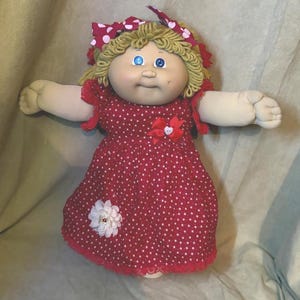 Könnte beinhalten: Eine Stoffpuppe mit blondem Wollhaar, blauen Augen und einem Gesicht mit Grübchen. Die Puppe trägt ein rotes Kleid mit weißen Punkten, das mit einer roten Schleife mit weißem Herzknopf an der Brust und einer weißen Stoffblume am Saum verziert ist.
