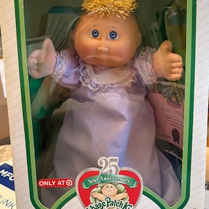 May include: A Cabbage Patch Kids Preemie doll in its original box. The doll has blue eyes, a light complexion, and a tuft of yellow hair. It wears a lavender dress with a white lace collar. The box features a 25th-anniversary logo and the text "I'm One of a Kind."