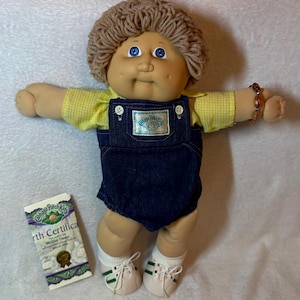 Può includere: Una bambola Cabbage Patch Kids con capelli in lana marrone chiaro e occhi blu, che indossa una camicia a quadretti gialla sotto una salopette in denim blu scuro con una toppa con logo. La bambola ha calzini e scarpe da ginnastica bianche a righe verdi.