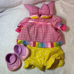May include: A doll outfit set featuring a pink plaid dress with a bow-shaped hood, yellow polka dot shorts, and purple shoes. The dress has rainbow trim and the shoes are approximately 2 inches long.