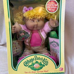 May include: A Cabbage Patch Kids doll with yellow yarn hair, brown eyes, and a pink bow, wearing a pink textured jacket over a pink shirt and tan pants. The doll is in a box with the text Includes Over 60 Fun CPK Stickers For You! and One of a Kind, And Just Mine!