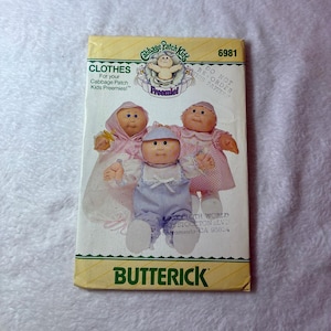 May include: A Butterick sewing pattern envelope for Cabbage Patch Kids Preemie clothes, number 6981. The front features illustrations of three dolls wearing various outfits, including a pink hooded gown, a blue striped romper, and a pink polka dot dress.