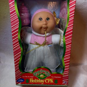 May include: A Cabbage Patch Kids Holiday CPK doll in a box, featuring a doll with a pink hat, a gold bow, and a white dress with a pink bodice. The doll has green eyes and is accompanied by a pink bottle. The packaging includes red and white stripes.