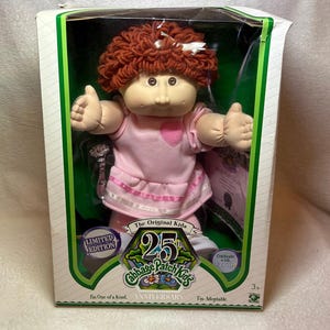 Może przedstawiać: Lalka Cabbage Patch Kids z okazji 25-lecia z włosami z kasztanowej włóczki i białą kokardką, ubrana w różową sukienkę z aplikacją w kształcie serca. Lalka znajduje się w pudełku z napisem 25th Anniversary Cabbage Patch Kids i Limited Edition.