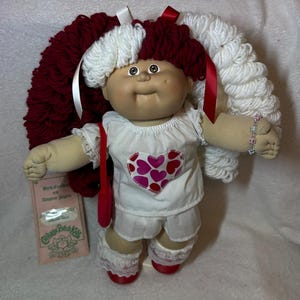 May include: A Cabbage Patch Kids doll with yarn hair in alternating sections of deep red and white, tied with ribbons. The doll wears a white top with a heart pattern, white shorts, and lace-trimmed socks. A tag labeled Birth Certificate is included.