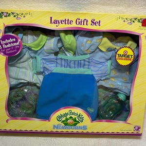 May include: A Cabbage Patch Kids Newborns Layette Gift Set in a yellow box. The set includes three doll fashions, a blue striped romper, a light blue patterned outfit, and a green bottle. The packaging features text including Layette Gift Set.